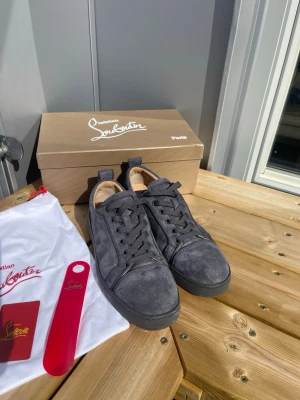 Gråa Louboutin sneakers i mocka - Snygga gråa sneakers från Christian Louboutin i mjuk mocka med klassisk röd sula. Skorna har lågt skaft, svarta snören och diskreta detaljer. Perfekta för dig som vill ha något exklusivt och stilrent till din outfit.