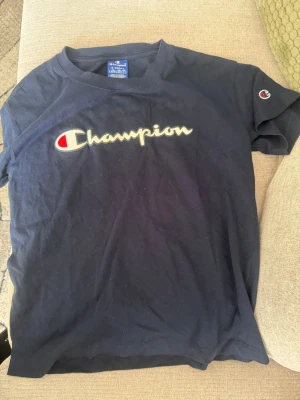 Mörkblå Champion t-shirt - Snygg mörkblå t-shirt från Champion med det klassiska logotrycket över bröstet och en liten logga på ärmen. Tillverkad i mjuk bomull och har en normal passform med korta ärmar. Perfekt för en avslappnad och sportig stil.