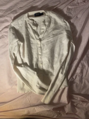 Vit cardigan från Polo Ralph Lauren - Klassisk vit cardigan från Polo Ralph Lauren med rund halsringning och knäppning framtill. Tröjan har långa ärmar och diskret broderad logga på bröstet. Tillverkad i mjukt material som känns skönt mot huden.