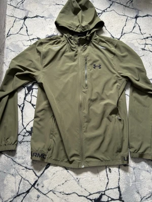 Olivgrön vindjacka från Under Armour - Snygg olivgrön vindjacka från Under Armour med huva, hel dragkedja och bröstficka med dragkedja. Jackan har elastiska muddar vid ärmsluten och diskret logga på bröstet. Perfekt för blåsiga dagar och sportiga outfits. Får original grejerna med