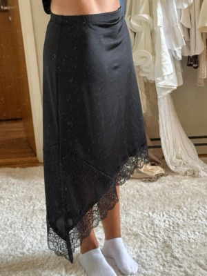 Svart långkjol med spetsdetalj - Säljer en svart långkjol med asymmetrisk fåll och snygg spetskant längst ner. Kjolen har en enkel, stilren design och är gjord i ett mjukt, lätt material som faller snyggt. Ny med prislapp.