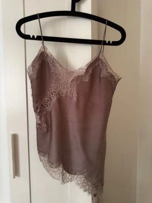 Linne i satin med spetsdetaljer - Säljer ett elegant linne i mjuk, skimrande taupefärgad satin med tunna axelband och vackra spetsdetaljer längs kanterna. Perfekt för dig som gillar en feminin och lyxig känsla. Materialet känns svalt mot huden och spetsen ger en romantisk touch.