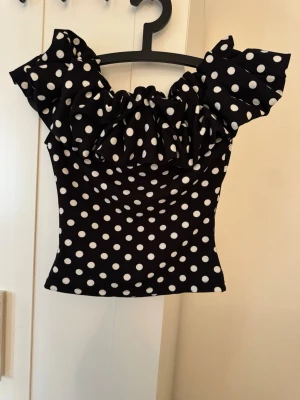 Svart prickig volangtopp - Säljer en svart topp med vita prickar och stora volanger över axlarna. Toppen är figurnära och har en offshoulder-design som ger en trendig look. Perfekt för dig som vill sticka ut med ett coolt mönster och snygga detaljer.