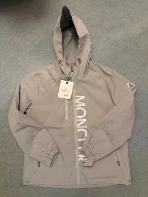 Ljusgrå vindjacka från Moncler - Snygg ljusgrå vindjacka från Moncler med huva och dragkedja framtill. Stor logga tryckt längs dragkedjan och klassisk Moncler-patch på ärmen. Jackan har två fickor med dragkedja och justerbar nederkant. Perfekt för blåsiga dagar. Storlek L men passar även i M och även i XL
