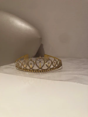 Guldigt diadem med strass och hjärtan - Snyggt diadem i guldfärgad metall dekorerat med gnistrande strass och hjärtformade detaljer. Diademet har kammar på sidorna för att sitta stadigt i håret och ger en elegant och lyxig känsla till din look.