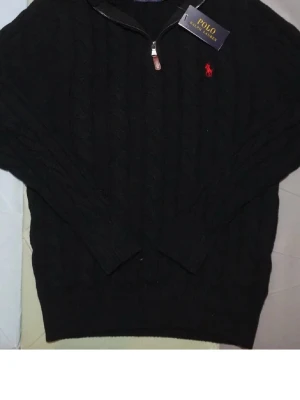 Svart quarter zip Polo Ralph Lauren (pris kan diskuteras) - Kabelstickad svart tröja från Polo Ralph Lauren med halv dragkedja och röd broderad logga på bröstet. Tröjan har långa ärmar, ribbade muddar och klassisk passform. Perfekt för dig som gillar stilrena och tidlösa plagg.