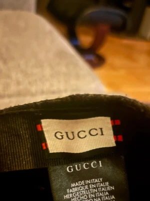 Svart keps från Gucci med logga.  - Snygg svart keps från Gucci med diskret GG-mönster över hela kepsen och klassiska gröna och röda ränder på sidan. Tillverkad i Italien och har justerbar rem bak. Perfekt accessoar för dig som vill ha en exklusiv och trendig look. Äkta med mycket bra kvalité. Kan ha den när och vart som helst.