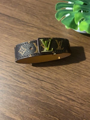 Armband - Snyggt armband i klassiskt brunt monogrammönster med guldfärgat spänne. Tillverkat i läder med tryckt logga och ikoniska detaljer. Perfekt accessoar för dig som gillar lyxig och tidlös stil.