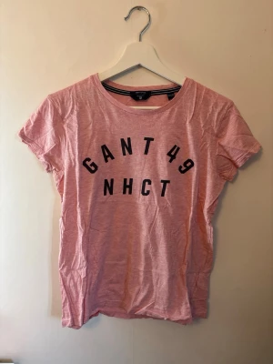 Rosa GANT t-shirt  - Säljer en rosa t-shirt från GANT med svart tryck 'GANT 49 NHCT' på bröstet. Klassisk rund halsringning och korta ärmar. Tillverkad i mjuk bomull som känns skön mot huden. Perfekt för en avslappnad och sportig stil.