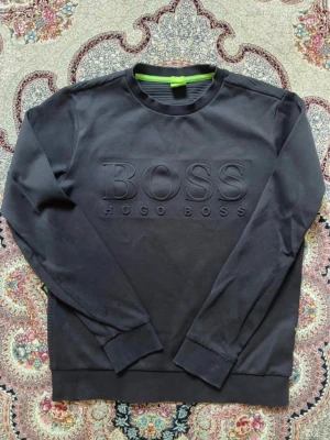 Svart sweatshirt från Hugo Boss - Stilren svart sweatshirt från Hugo Boss med stort präglat BOSS-tryck på bröstet. Tröjan har rund hals, ribbade muddar och är i slim fit-modell. Perfekt för dig som gillar en clean och modern look.