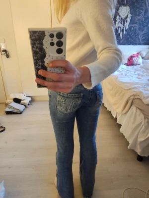 Blå raka jeans med detaljerad ficka - Säljer ett par blå jeans med raka ben och snygga detaljer på bakfickorna. Jeansen har ljus tvätt och dekorativa sömmar samt nitar på fickorna. Klassisk femficksmodell i mjukt denimtyg, perfekta till vardagslooken.