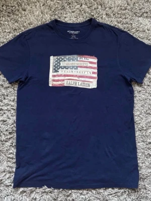 Mörkblå t-shirt Ralph Lauren flagga - Snygg mörkblå t-shirt från Ralph Lauren Denim & Supply med amerikansk flagga och vintage-tryck på bröstet. Tillverkad i 100% bomull och har klassisk rund hals och korta ärmar. Perfekt för en avslappnad och cool stil.