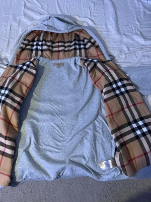 Grå zip från Burberry - Snygg grå jacka från Burberry i storlek XL med det klassiska rutiga Burberry-mönstret på insidan. Jackan har huva och är tillverkad i mjukt material, perfekt för dig som vill ha en stilren och tidlös look. XL men sitter bra om du har L