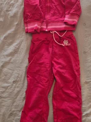 Rosa mjukisdress med dragkedja - Säljer en rosa mjukisdress med dragkedja och ribbade muddar. Byxorna har dragsko i midjan och ett broderat mönster på ena sidan. Perfekt för chill dagar hemma eller som loungewear. Materialet är mjukt och skönt mot huden. i storlek 86