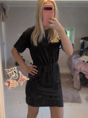Svart glittrig kortklänning - En svart kortklänning med glittrigt tyg och resår i midjan. Klänningen har rund halsringning och korta ärmar, vilket ger en enkel men snygg look. Perfekt för dig som vill ha något stilrent men ändå lite extra. Perfekt för nyår