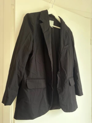 Svart oversized kavaj med fickor - Stilren svart kavaj i oversized modell med klassisk krage och en knapp framtill. Två framfickor med lock och en bröstficka. Perfekt för en trendig och avslappnad look. 