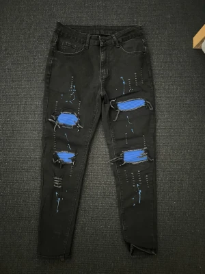 Svarta ripped jeans med blå detaljer - Säljer ett par svarta skinny jeans med stora slitningar och blå tygpatchar under hålen. Jeansen har även blå färgstänk och råa kanter vid bensluten. Perfekt för dig som gillar en edgy och trendig stil.