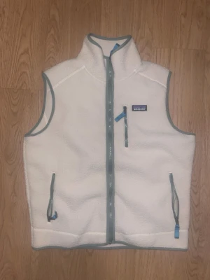 Vit fleeceväst från Patagonia - Snygg vit fleeceväst från Patagonia med grå detaljer och dragkedjor. Västen har hög krage, tre fickor med dragkedja och mjukt, fluffigt material som håller värmen. Perfekt för lager-på-lager och outdoor-vibe.