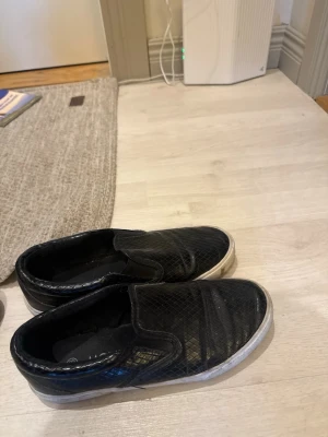 Svarta slip-on sneakers med mönster - Svarta slip-on sneakers med vit sula och diskret rutmönster på ovandelen. Skorna har en låg profil och elastiska paneler på sidorna för enkel på- och avtagning. Perfekta för en avslappnad och stilren look.
