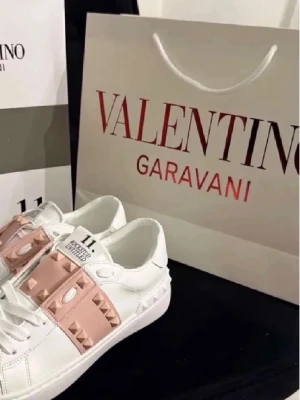 Valentino Rockstud Untitled sneakers - Snygga vita sneakers från Valentino med ljusrosa band och ikoniska nitar över vristen. Skorna har en klassisk låg siluett och är tillverkade i läder med coola detaljer som ger en lyxig vibe.