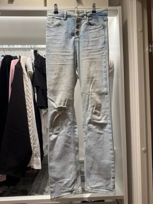 Ljusblå raka jeans med lite utsvängning  - Säljer ett par ljusblå jeans med raka ben och hög midja. Jeansen har slitning och hål på ena knät samt knappar i gylfen. Klassisk femficksmodell i ett tvättat denimtyg. Perfekta för en avslappnad och trendig look. Midjemått 31,5cm, midjan till skrevet 27 cm, innrebenslängd 70cm 