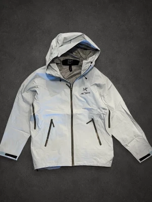 Ljusblå  regnjacka från Arc'teryx - Snygg ljusblå regnjacka från Arc'teryx med huva och hel dragkedja framtill. Jackan har två snedställda fickor med dragkedja och justerbara muddar vid ärmsluten. Tillverkad i ett tunt, vattentätt material som passar perfekt för regniga dagar.