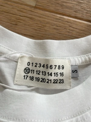 Maison Margiela vit t-shirt med tryck - Vit t-shirt från Maison Margiela med ikoniskt siffertryck och stor logga på framsidan. Klassisk rund hals och korta ärmar. Tillverkad i mjuk bomull för en skön och avslappnad look.
