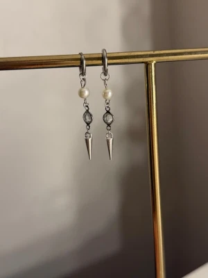 Silvriga örhängen med pärlor och spikes - Snygga örhängen i silverfärg med vita pärlor, små runda stenar och coola spetsdetaljer längst ner. Perfekta för att ge din look en edgy touch. Hängande modell som passar till många stilar.