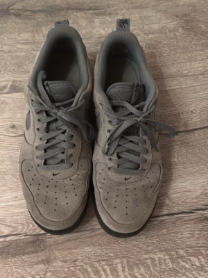 Grå Nike Air Force 1 sneakers - Säljer ett par grå Nike Air Force 1 sneakers i mocka med mörkgrå sula och matchande grå skosnören. Swoosh-loggan på sidan har ett unikt mönster och skon har klassiska perforeringar på tån för ventilation. Perfekt för dig som gillar stilrena och sportiga sneakers.