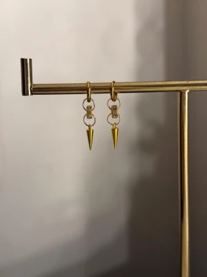 Guldpläterade örhängen med spetsdetalj - Säljer ett par trendiga örhängen i guldpläterad metall med små ringar och en spetsig hängande detalj. Örhängena har en minimalistisk men edgy design som sticker ut. Perfekta för dig som gillar unika accessoarer och vill addera lite attityd till din stil.