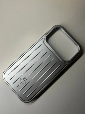 iPhone 17 PRO skal rimowa silver  - Rimowa mobil skal till iPhone 17 PRO. Skalets design är stilren och modernt minimalistisk, med ett stort kamerauttag och en diskret logotyp. Materialet känns robust och ger ett exklusivt intryck.