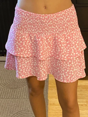 Rosa volangkjol med blommönster - Söt rosa kortkjol med vita små blommor och volangdetaljer. Storlek L, men passar även mig i storlek XS/S pga midjan är i smock!