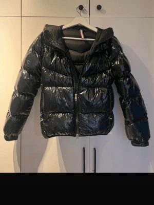 Svart glansig Colmar jacka   - Säljer en svart, glansig Colmar vinterjacka med huva och dragkedja framtill. Jackan har breda, vadderade paneler och två sidofickor. Materialet är syntetiskt och ger en riktigt cool look. Perfekt för kalla vinterdagar när du vill hålla dig varm och se trendig ut.