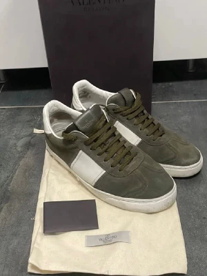 Olivgröna sneakers från Valentino - Snygga olivgröna sneakers från Valentino med vita detaljer och matchande olivgröna skosnören. Skorna har en låg profil och är tillverkade i mocka och läder. Klassisk design med vit sula och diskret Valentino-logga på plösen . Några nitar är borta samt så har skorna blivit limmade där back då skon gått upp lite och de  lite lite smutsiga är de