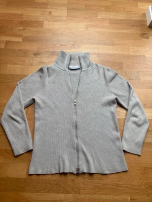Ziptröja Ljung - Snygg beige ribbad tröja med dragkedja hela vägen och hög krage. Tröjan har långa ärmar och är tillverkad i ett mjukt, stretchigt material som sitter skönt på kroppen. Perfekt för lager på lager och enkel att matcha med olika outfits.