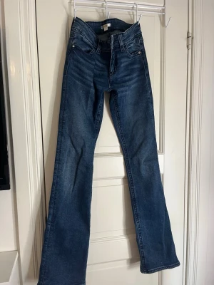 Blå bootcut jeans med ganska låg midja  - Säljer ett par klassiska blå bootcut jeans med relativt låg midja och fyra fickor. Jeansen har en snygg tvätt och lätt utsvängda ben som ger en retrovibe. Perfekta att styla med sneakers eller boots. Materialet är mjukt denim i bomull.