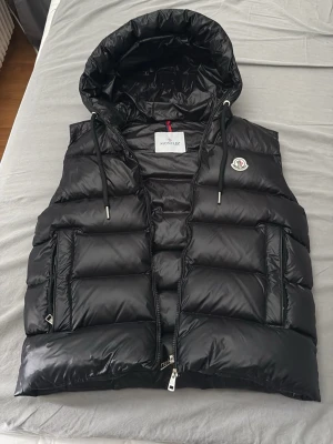 Svart dunväst med huva från Moncler - Snygg svart dunväst från Moncler med glansig finish och stor huva. Västen har två stora fickor med dragkedja, dragsko i huvan och klassisk Moncler-logga på bröstet. Insidan har ett unikt serietryck. Perfekt för lager på lager och streetwear.