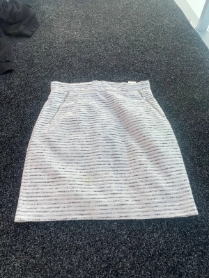 Vit och grå randig kortkjol - Säljer en vit och grå kortkjol med tunna horisontella ränder. Kjolen har fickor framtill och en diskret dragkedja i sidan. Perfekt för dig som gillar enkel men stilren design.