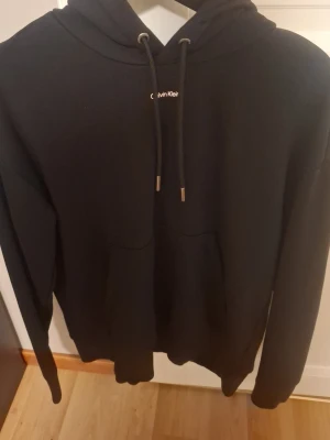 Svart hoodie från Calvin Klein - Säljer en svart hoodie från Calvin Klein med klassisk huva och dragsnören. Hoodien har en diskret logga på bröstet och en stor magficka framtill. Tillverkad i mjukt material och har långa ärmar. Perfekt för en avslappnad och stilren look. Använd inte så mycket alls då jag ej gillade den