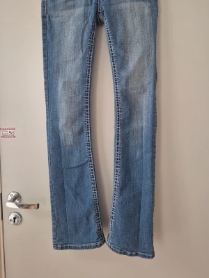 Blå bootcut jeans med broderade fickor - Säljer ett par klassiska blå bootcut jeans med snygga broderade detaljer på bakfickorna. Jeansen har normal midja och är tillverkade i ett mjukt denimtyg som ger en avslappnad look. Perfekta för dig som gillar en tidlös och trendig stil. Kolla noga genom bimder så ser du att på höger sida finns det ett litet hål. Lätt att sy. Hund finns i hemmet. 
