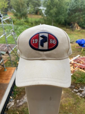Peak performance keps - Stilren beige keps med böjd skärm och broderad patch framtill med siffrorna 1986 och ett blått emblem. Kepsen har klassisk passform och är tillverkad i bomullstyg. Perfekt accessoar för en avslappnad look.