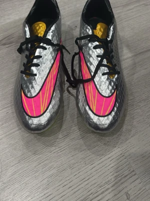 Nike Hypervenom Phantom - Snygga fotbollsskor i silver med stora rosa och gula detaljer på sidorna. Skorna har svart snörning och en gul plös. Ovandelen har en texturerad yta för bättre bollkontroll och en modern, sportig look. Perfekta för dig som vill sticka ut på planen.