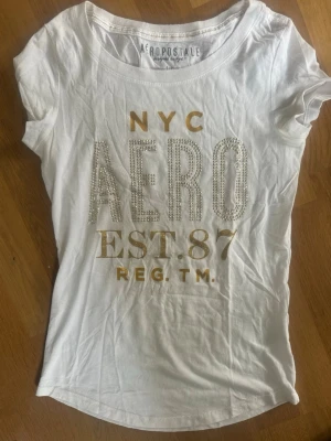 Vit t-shirt från Aeropostale NYC - Vit t-shirt från Aeropostale med guldfärgad text och pärldetaljer på bröstet. Trycket har texten 'NYC AERO EST. 87 REG. TM.' T-shirten är kortärmad och har en klassisk rund hals. Materialet är en mix av bomull och polyester. Använd men fungerar väl.