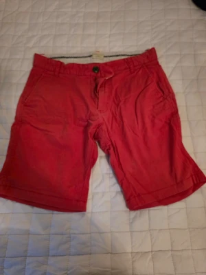Röda shorts från Selected Homme - Snygga röda shorts från Selected Homme med klassisk passform. Shortsen har knapp och dragkedja framtill samt sidofickor. Perfekta för varma dagar och enkla att matcha med t-shirt eller skjorta.