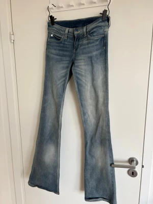 Blå bootcut jeans med slitningar - Snygga blå bootcut jeans med lätt slitna detaljer och klassisk femficksdesign. Jeansen har dragkedjegylf och mässingsfärgad knapp framtill. Perfekta för en avslappnad och trendig look.