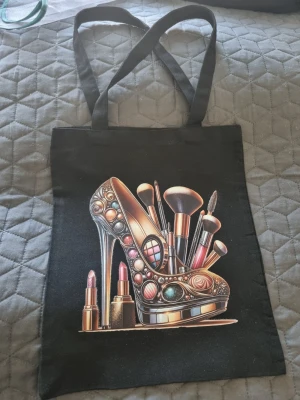 Svart shoppingväska med sminkmotiv - Cool svart shoppingväska i tyg med tryck av en glittrig högklackad sko fylld med sminkborstar, läppstift och sminkdetaljer. Perfekt för dig som älskar makeup och vill ha en unik accessoar. Väskan har långa handtag och rymlig insida. Hangjord av mig 🌺Oanvänd 