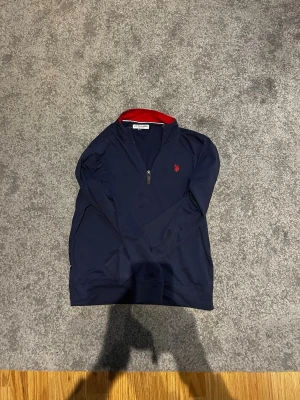 Marinblå halvzip från U.S. Polo Assn - Marinblå långärmad tröja från U.S. Polo Assn med halv dragkedja och röd broderad logga på bröstet. Kragen har en snygg röd insida som ger extra kontrast. Perfekt till jeans eller chinos för en clean och sportig stil.
