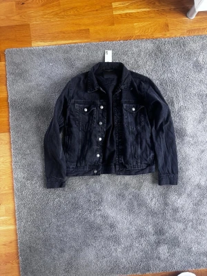Svart jeansjacka med silverknappar - Snygg svart jeansjacka med klassisk krage och silverfärgade knappar framtill. Två bröstfickor med lock och knapp samt raka ärmar. Perfekt att slänga över en hoodie eller t-shirt för en cool look.