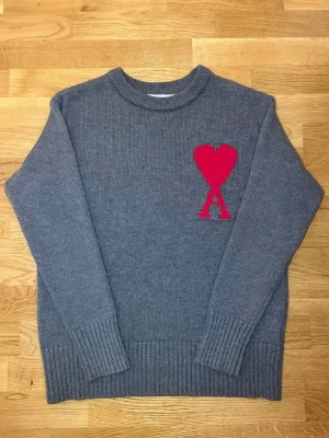 Grå stickad tröja med rött hjärta - Mysig grå stickad tröja med rund hals och ribbade muddar. På bröstet finns ett stort rött hjärta och bokstaven A, vilket ger en unik och trendig look. Perfekt för dig som vill ha något stilrent men ändå med en cool detalj.