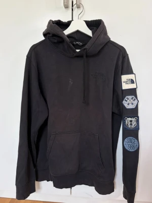 Svart hoodie från The North Face - Svart hoodie från The North Face med huva och känguruficka. Ärmen har flera patchar i blått och vitt, bland annat med logga och texten 'Since 1966'. Tillverkad i mjukt bomullsmaterial och har ribbade muddar vid ärmslut och nederkant. Har en fläck framtill och är lite solblekt